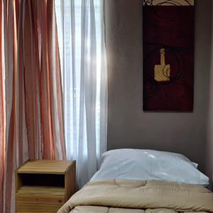 Habitación individual luminosa con cama, armario, mesa de luz, aire acondicionado y ventana grande en la residencia para mayores Casa Trapiche.